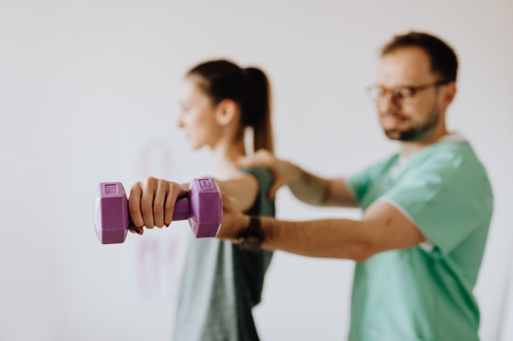 pexels-photo-4506112 Side view of professional orthopedist in uniform and eyewear helping fit woman reaching arm with dumbbell in doctor office on blurred background